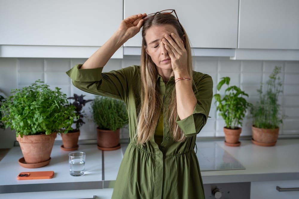 A woman in a green dress holds her glasses and rubs her eyes in the kitchen, signs of perimenopause tiredness. Various potted plants fill the background, while a glass of water sits on the counter.