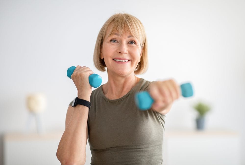 A person with short blonde hair exercises with blue dumbbells, wearing a grayish-green shirt and a smartwatch, against a soft-focus neutral background.