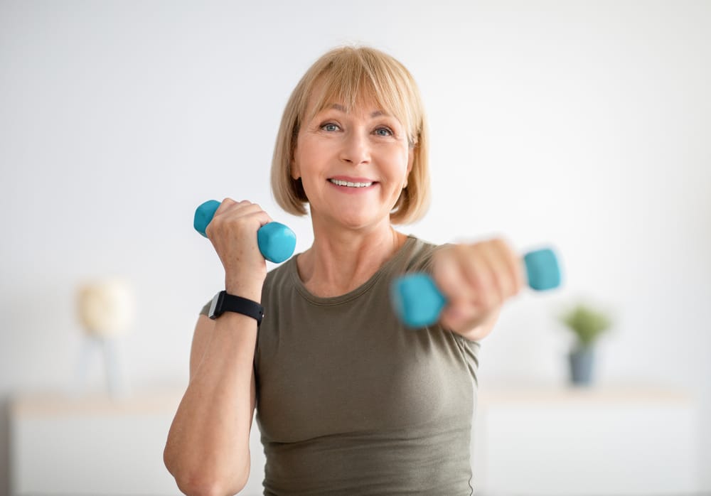 A person with short blonde hair exercises with blue dumbbells, wearing a grayish-green shirt and a smartwatch, against a soft-focus neutral background.